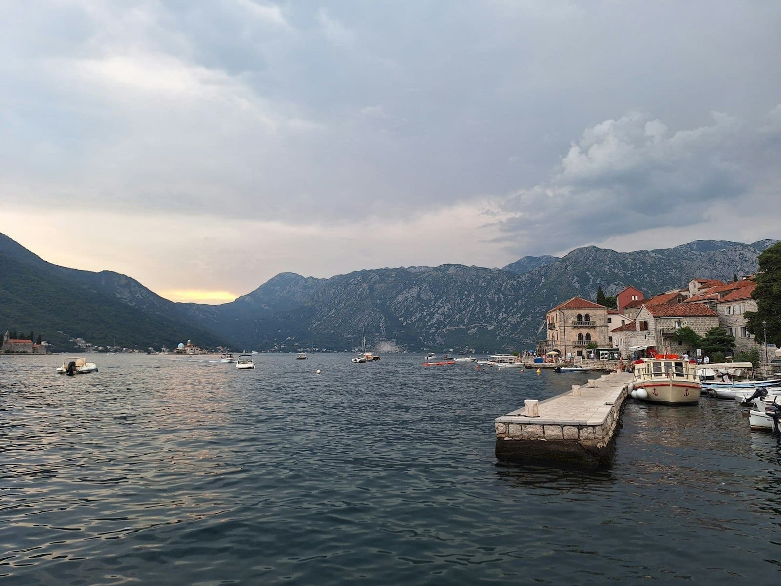 Vue panoramique du front de mer de la baie de Kotor avec de petits bateaux, un quai en pierre et des bâtiments historiques adossés aux montagnes au crépuscule.