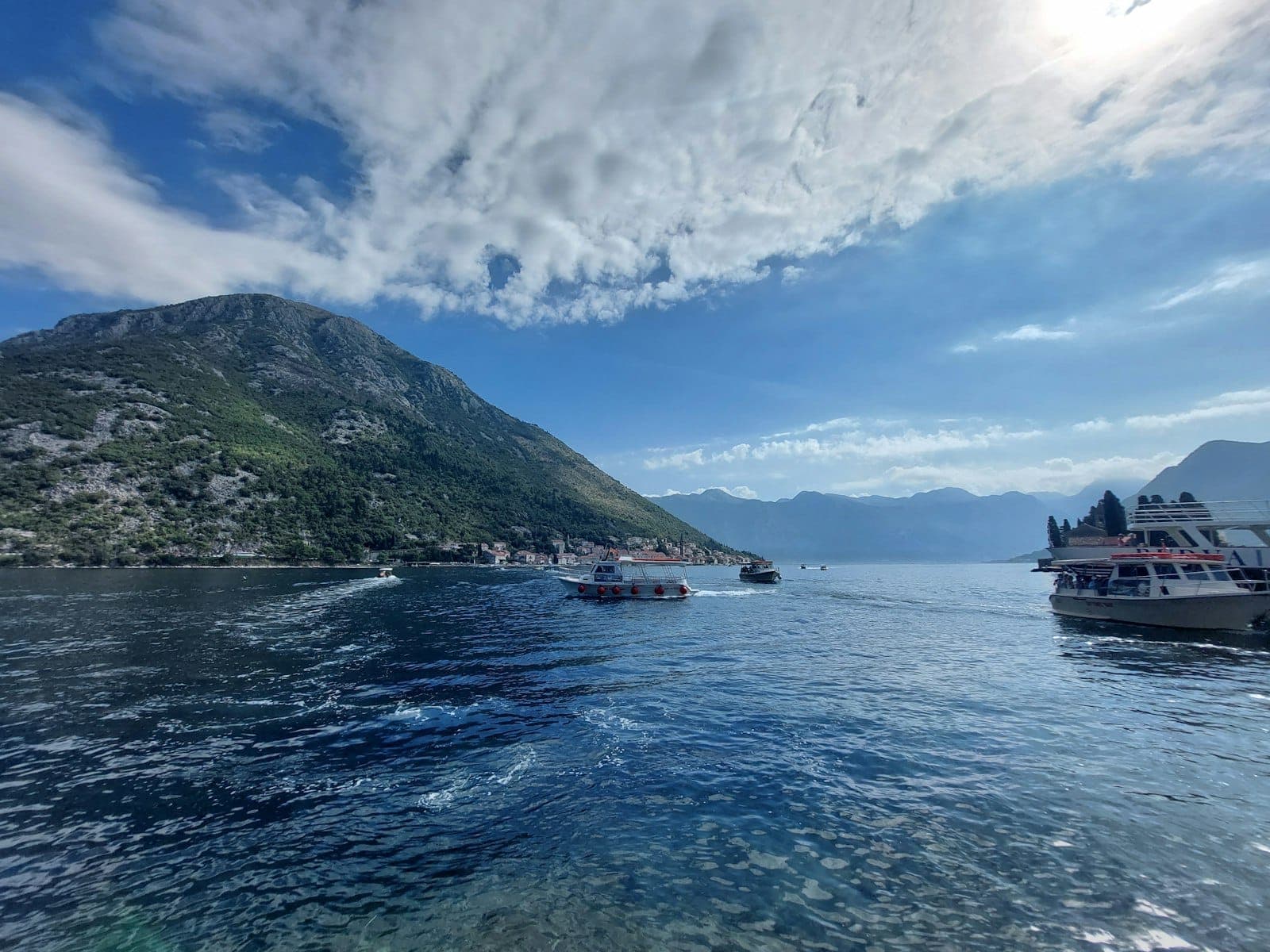 Vue panoramique du littoral de Kotor avec des bateaux sur une eau bleue et claire sous un ciel ensoleillé, des montagnes au loin et des reflets marins éclatants.