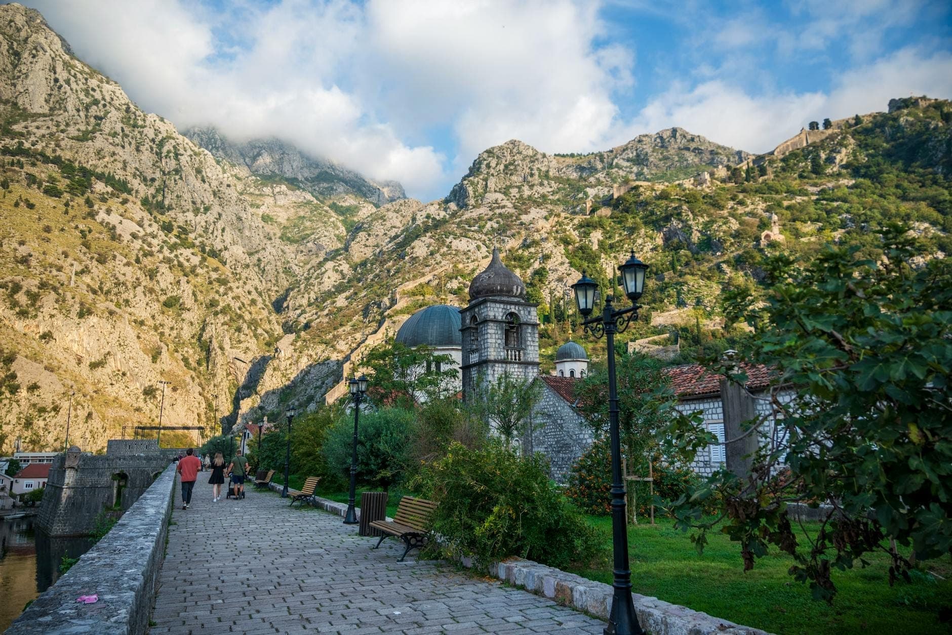 Promenade piétonne à Kotor avec des personnes marchant vers les montagnes, des bancs et de la verdure le long d'un chemin de pierre.