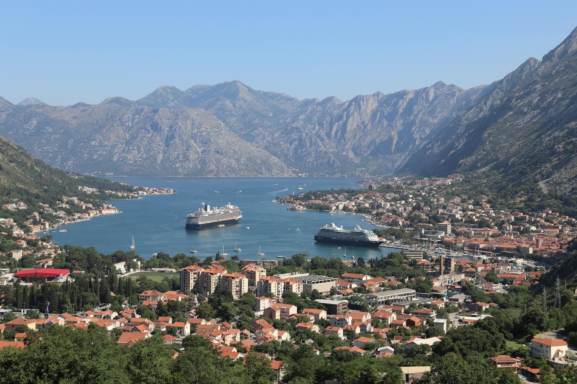 Vue panoramique ensoleillée de la Baie de Kotor avec des bateaux de croisière amarrés et un temps clair sur la ville et les montagnes.