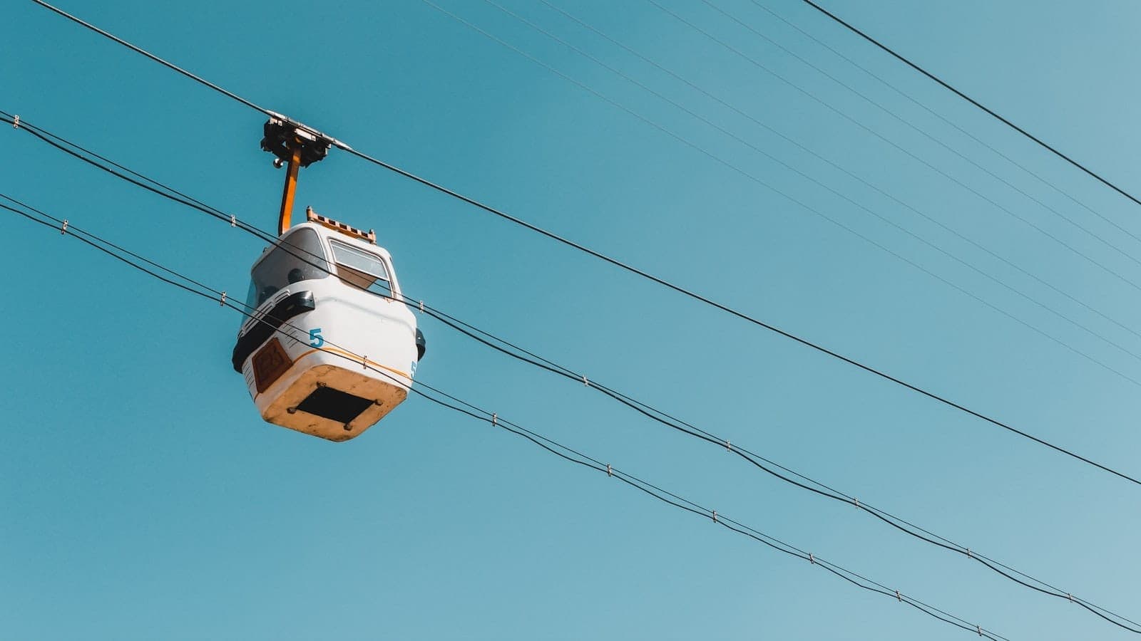 Cabine blanche du téléphérique de Kotor se déplaçant sur les câbles contre un ciel bleu dégagé, photographiée depuis le bas par une journée ensoleillée.