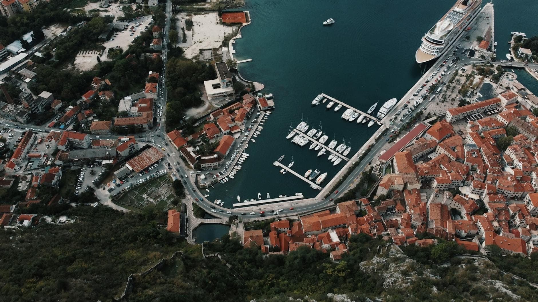 Vue aérienne du port de croisière de Kotor avec un grand navire de croisière amarré près de la vieille ville et la marina, entouré de bâtiments aux toits rouges.