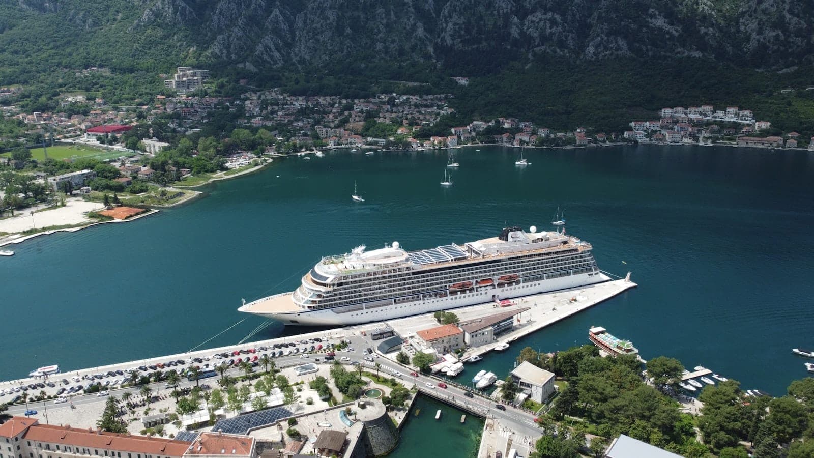 Vue aérienne d'un grand navire de croisière amarré au port de croisière de Kotor, entouré de collines verdoyantes, de bâtiments en bord de mer et d'eaux bleues calmes.