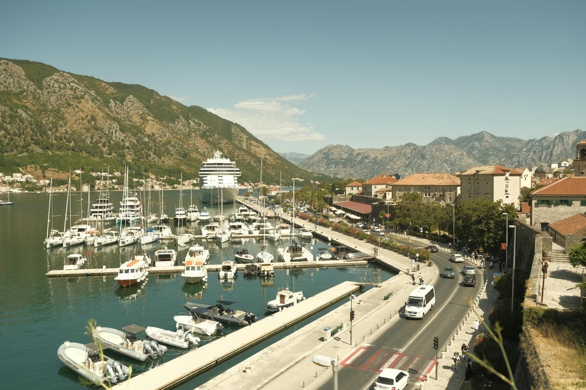 Une marina à Kotor bordée de yachts et de bateaux, avec une promenade en bord de mer, une route animée et des montagnes se découpant à l'arrière-plan sous un ciel dégagé.