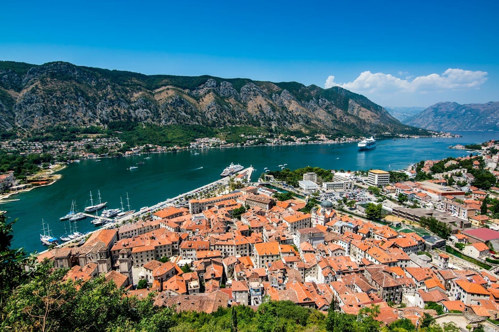 Vue panoramique de la vieille ville de Kotor avec ses toits de tuiles rouges encadrés par les remparts de la forteresse, la baie de Kotor et les montagnes environnantes sous un ciel d'un bleu éclatant.