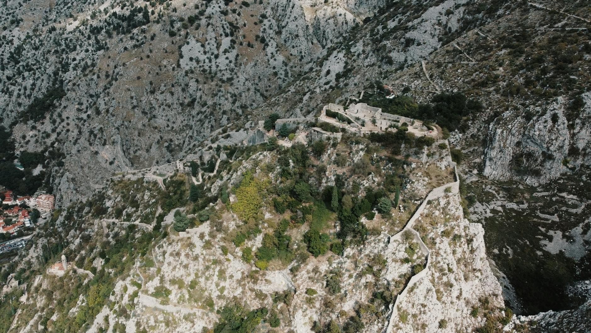 Vue aérienne des remparts de Kotor serpentant sur la flanc de montagne escarpé, montrant des fortifications et des marches en pierre au milieu d'un terrain rocheux.