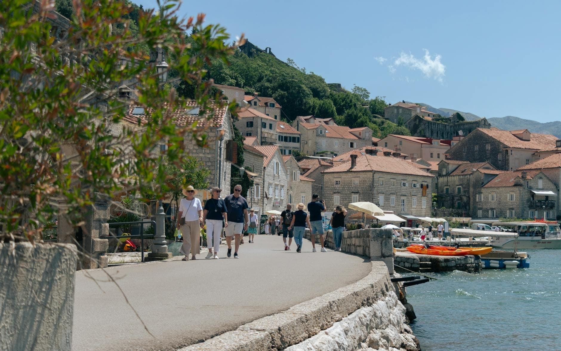 Des promeneurs déambulent sur la promenade en bord de mer de Kotor, avec des bâtiments en pierre, des bateaux et des montagnes en arrière-plan par une belle journée ensoleillée.