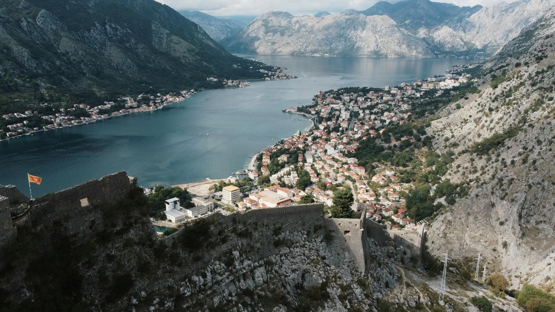 A stunning panoramic view over the Bay of Kotor, showing the walled old town below, surrounded by dramatic mountains and blue water under a partly cloudy sky.