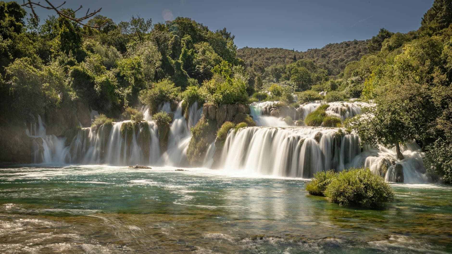 Weitwinkelpanorama der Kravica-Wasserfälle, die über Travertinfelsen in ein türkisfarbenes Becken stürzen, umgeben von üppig grünen Bäumen unter einem wolkenlosen Himmel.