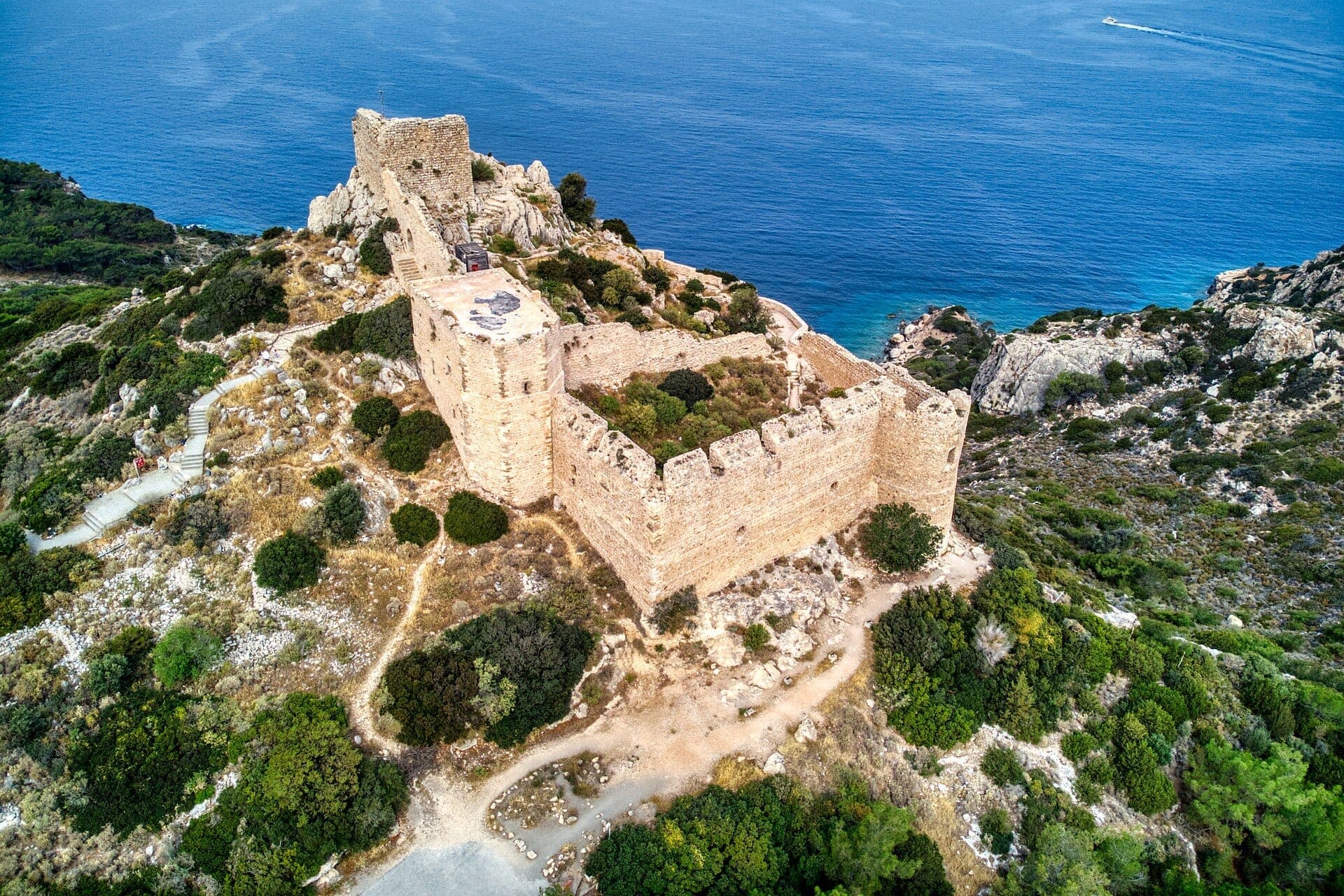 Vue aérienne des ruines du château de Kritinia au sommet d'une colline rocheuse, entourées d'une végétation sauvage et dominant le bleu profond de la mer Égée.