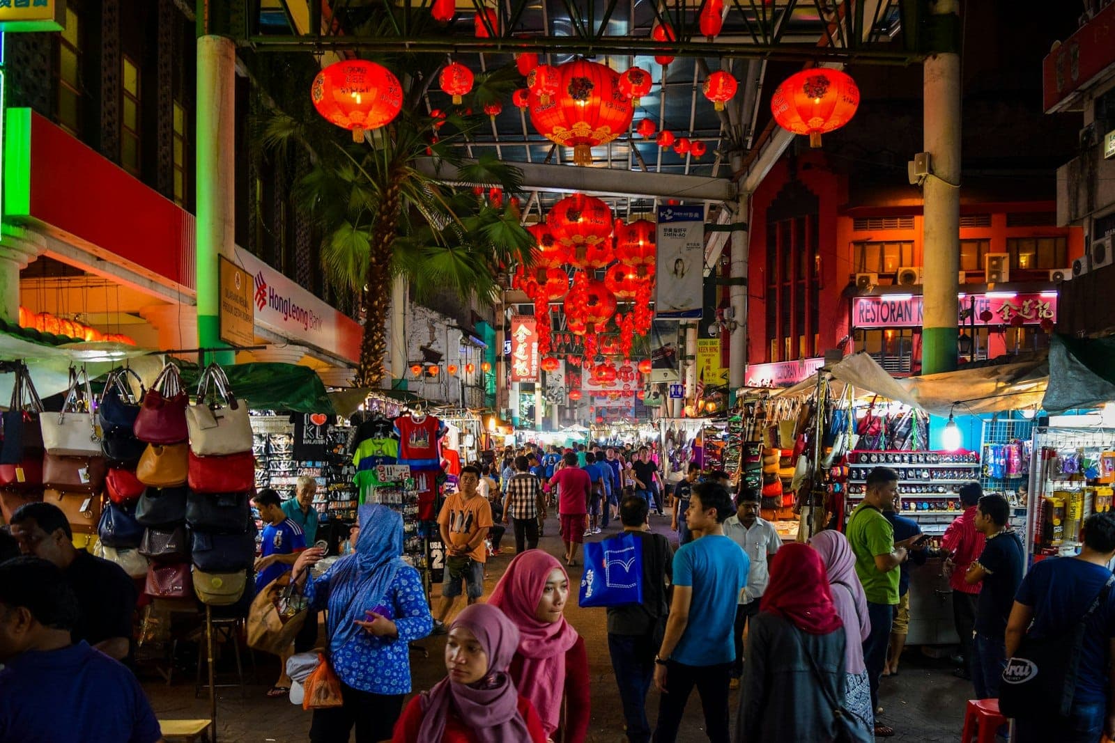 Einkäufer stöbern unter roten Laternen an lebhaften Straßenständen auf dem belebten Nachtmarkt von Kuala Lumpur, mit bunten Taschen und Kleidungsstücken auf beiden Seiten.