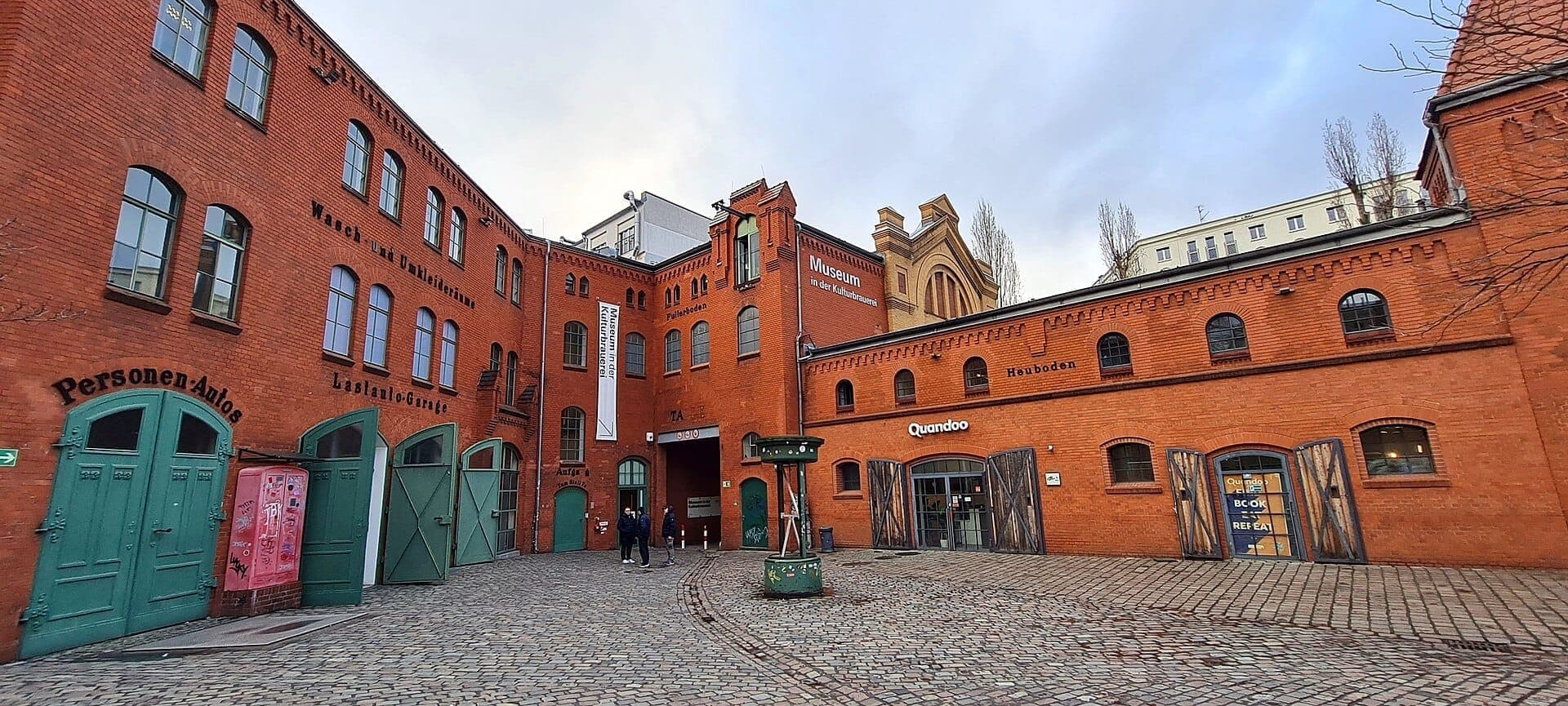 วิวอาคารอิฐแดงและลานหินกรวดของ Kulturbrauerei ในเบอร์ลิน พร้อมประตูสีเขียวและป้ายพิพิธภัณฑ์ที่มองเห็นได้ชัดเจน
