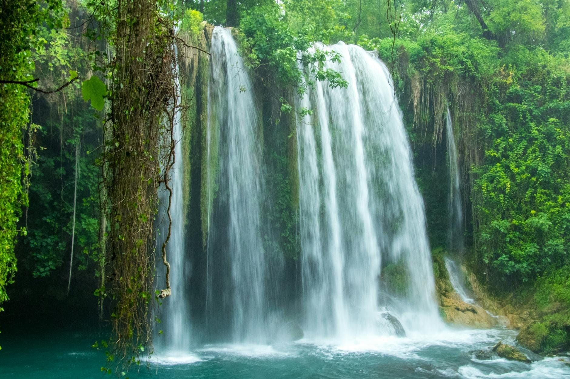Ein üppiger grüner Wald umgibt einen breiten, kaskadierenden Wasserfall, der in türkisfarbene Becken stürzt – Nebel und leuchtende Vegetation sorgen für eine frische, einladende Atmosphäre.