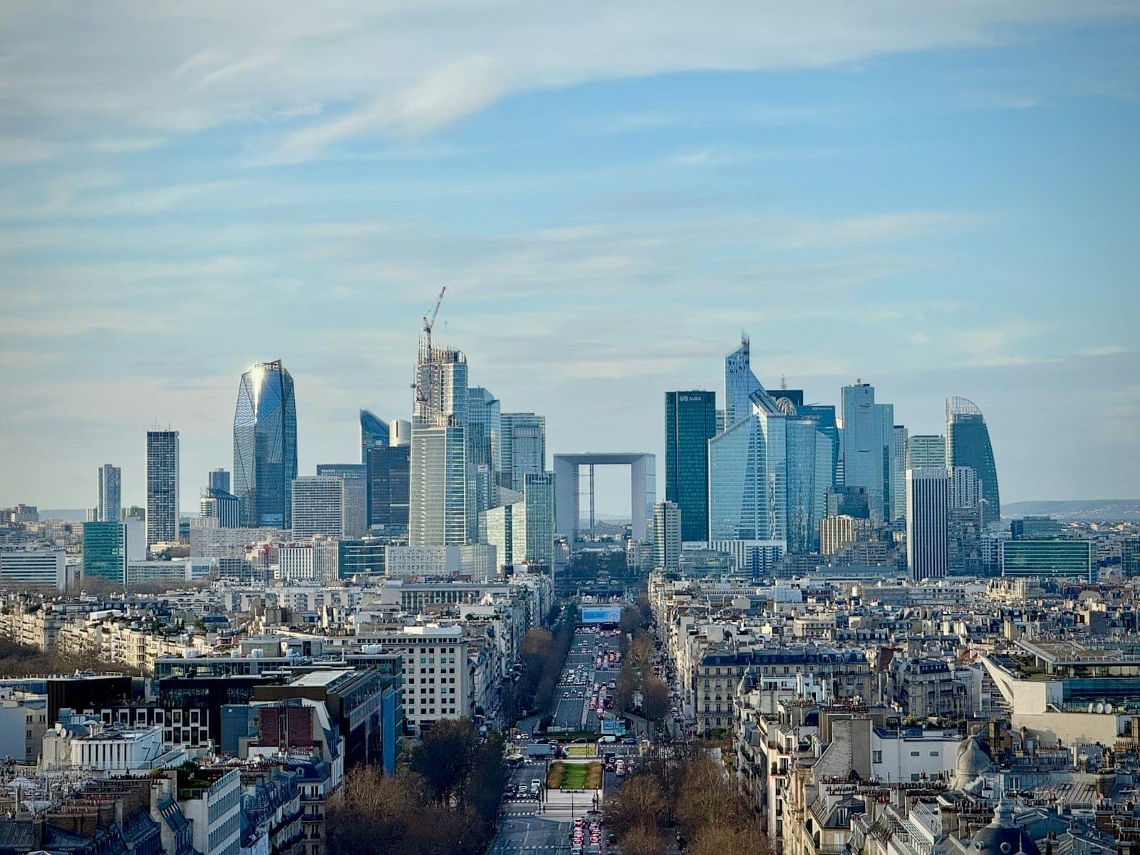 Weitläufiges Stadtpanorama von La Défense mit Glashochhäusern und der Grande Arche, aus der Entfernung über den Pariser Dächern unter blauem Himmel.