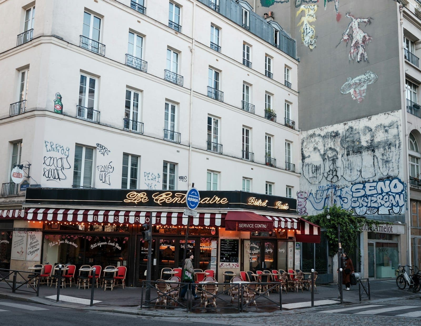 Parisian street corner with a classic café, outdoor tables, street art and graffiti on nearby building, in the vibrant 19th arrondissement.