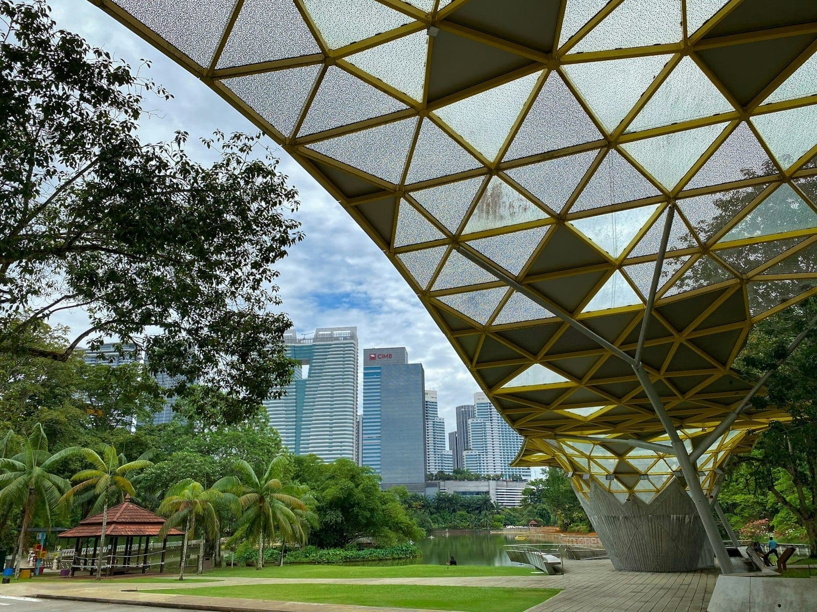 Kuala Lumpur Göl Bahçeleri geometrik gölgelik yürüyüş yolu, yemyeşil bitki örtüsü ve arka planda KL siluetiyle