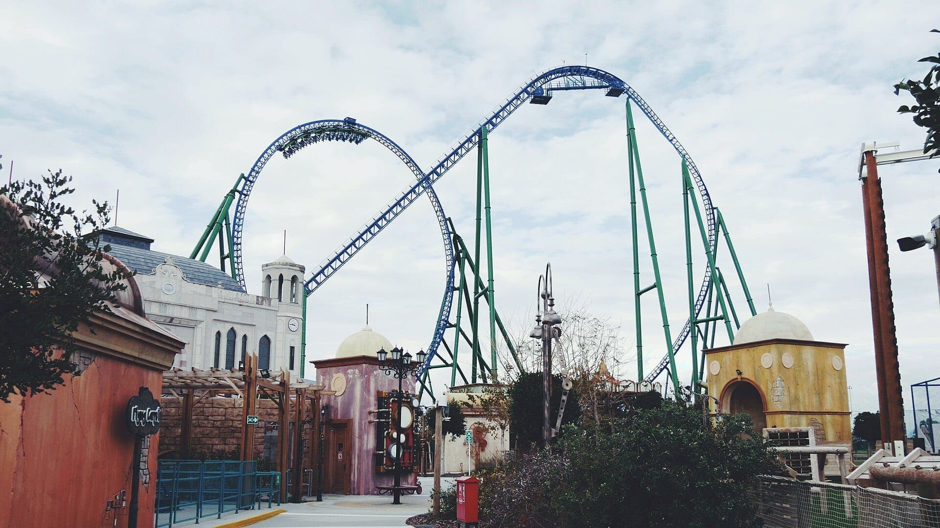 Hohes blau-grünes Achterbahngleis mit Loopings hinter kunstvoll gestalteten Themengebäuden im Land of Legends Theme Park in Antalya unter einem bewölkten Himmel.