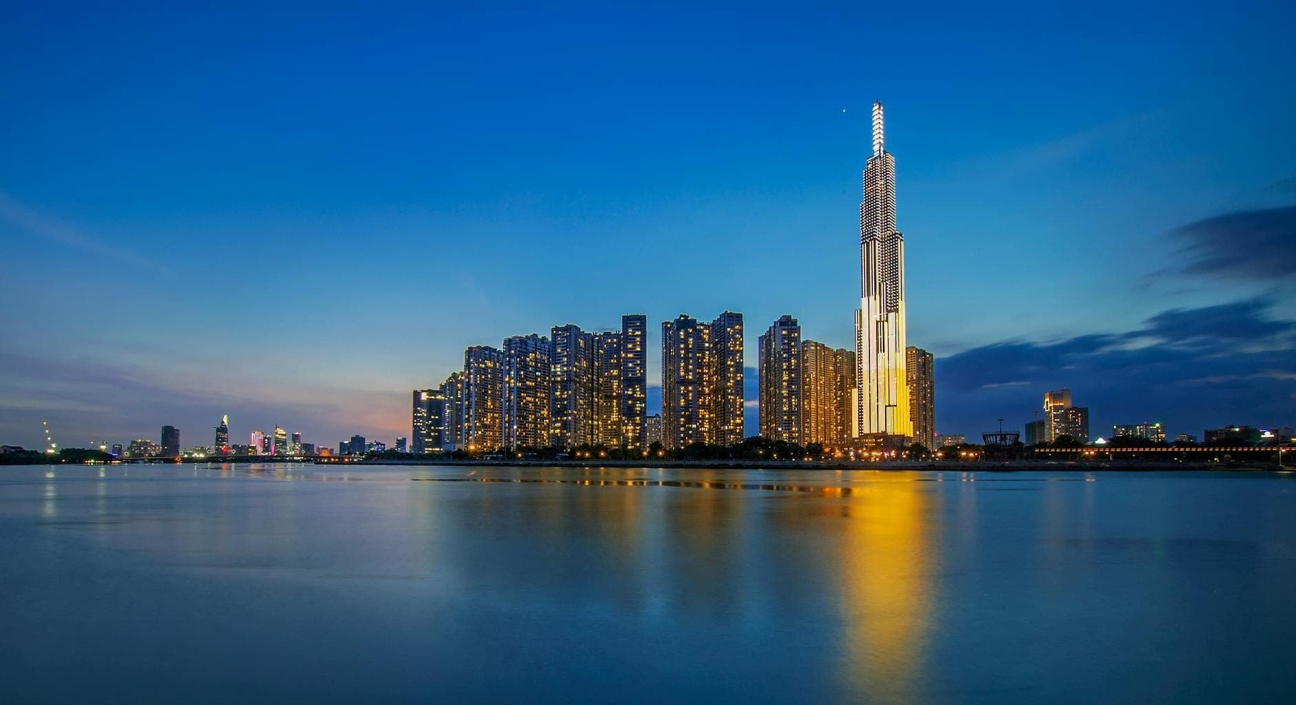 O Landmark 81 iluminado ao entardecer, dominando os edifícios ao redor e refletido nas águas calmas do rio sob um céu azul vibrante.