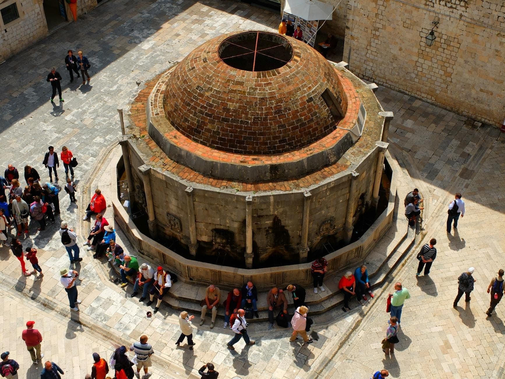 Luftaufnahme des Großen Onofrio-Brunnens in Dubrovnik mit Menschen, die um das kreisförmige Steinbauwerk sitzen und spazieren.