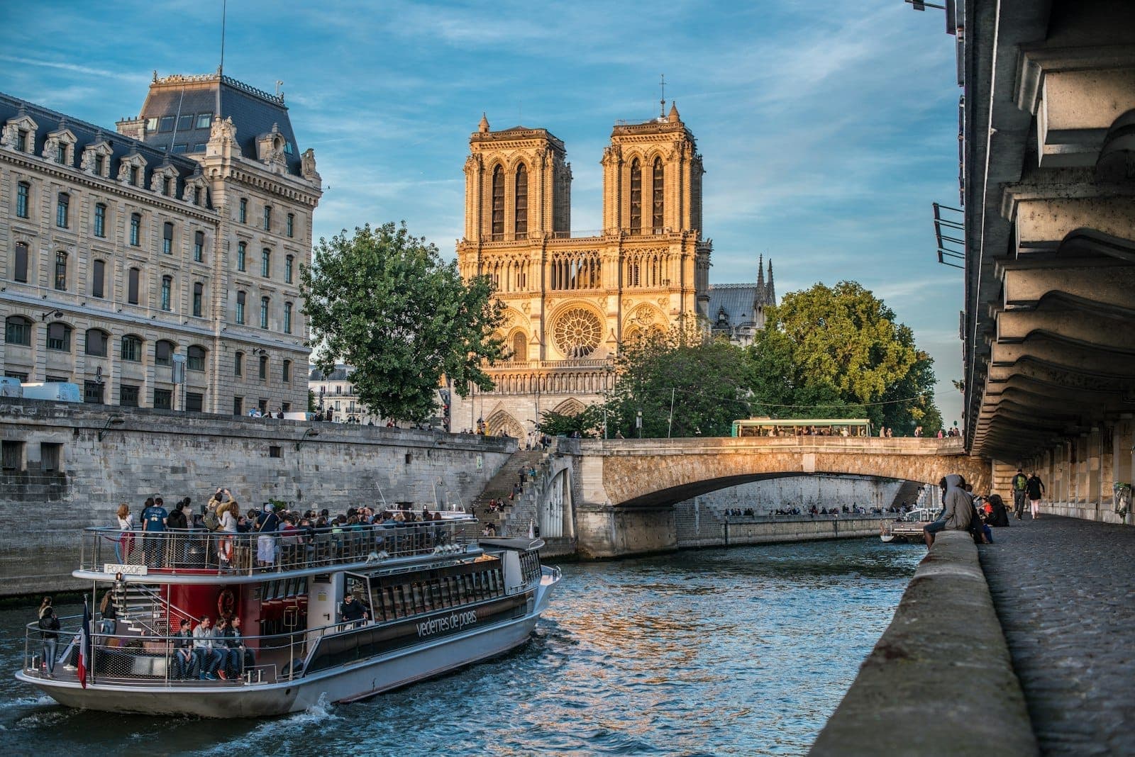 View of Notre-Dame Cathedral and the Seine River at golden hour, with a riverboat and pedestrians capturing the lively spirit of the Latin Quarter.