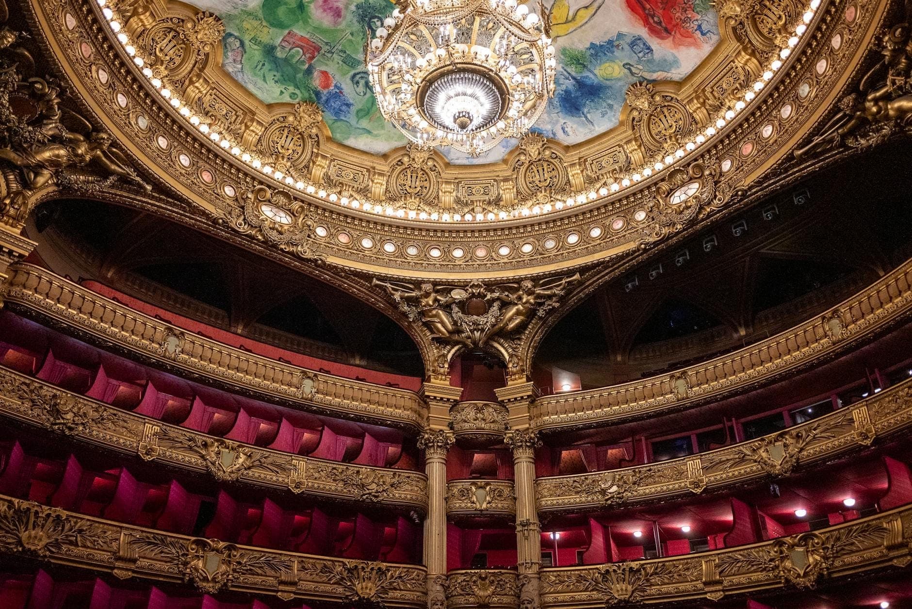 Opulent Art Deco theater interior with red velvet seats, ornate gold balconies, and a grand colorful ceiling with a sparkling chandelier.