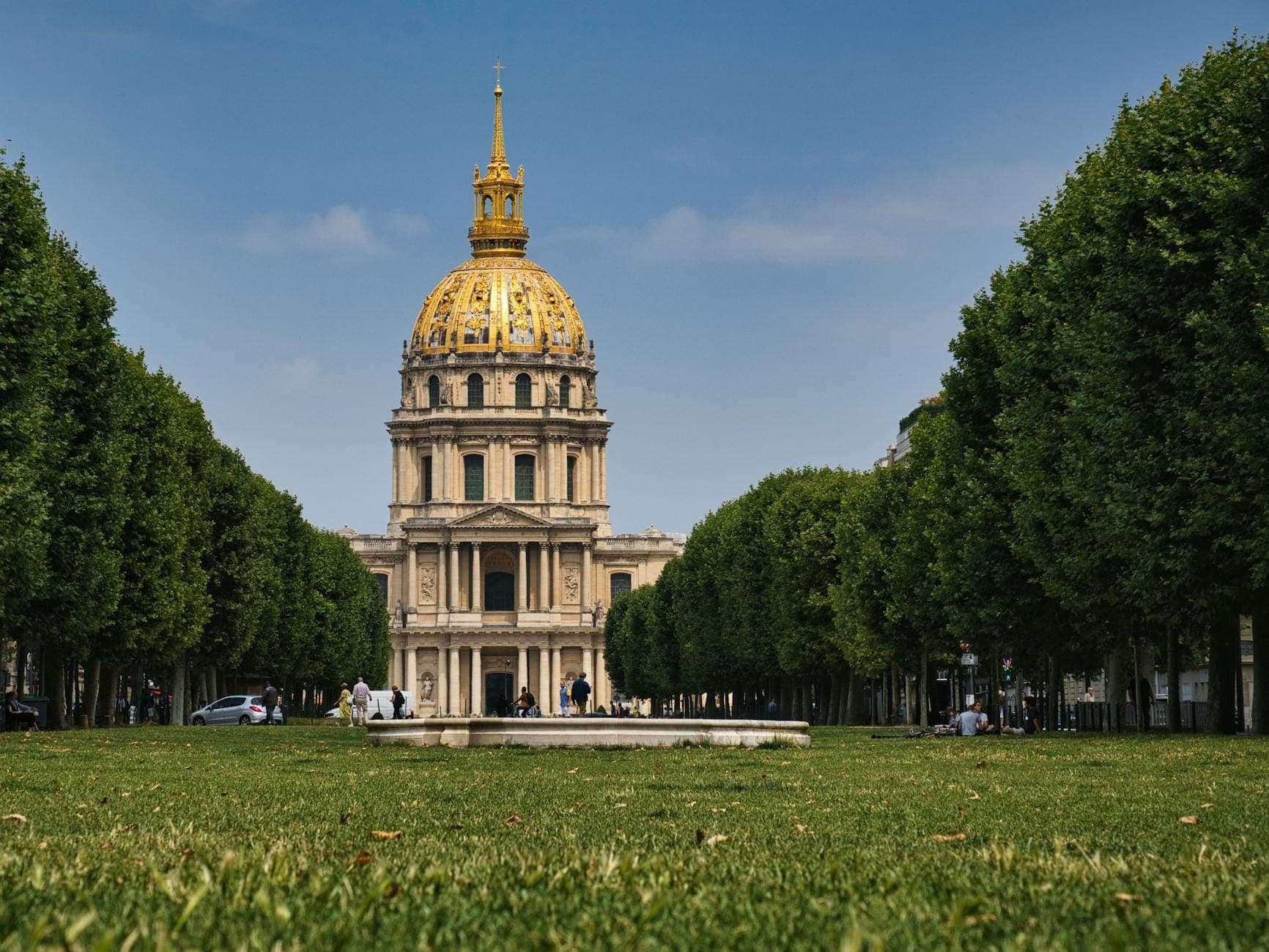 Front view of Les Invalides golden dome in Paris framed by a lush green lawn and leafy trees under a bright blue sky.