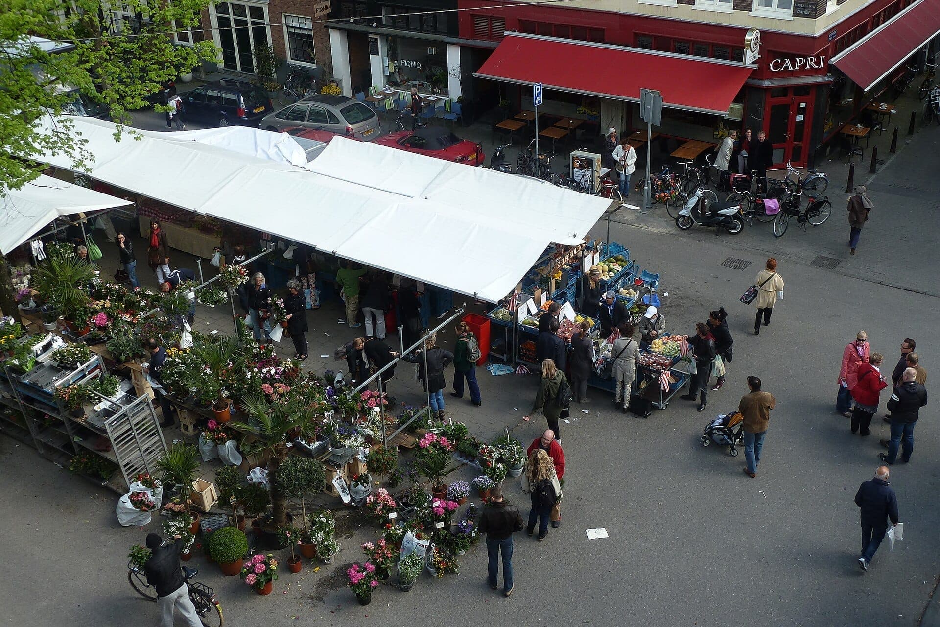 Luftaufnahme des Lindengracht-Markts mit Blumenständen, Produktständen und Menschen, die unter weißen Markisen an einer belebten Straßenecke schlendern.