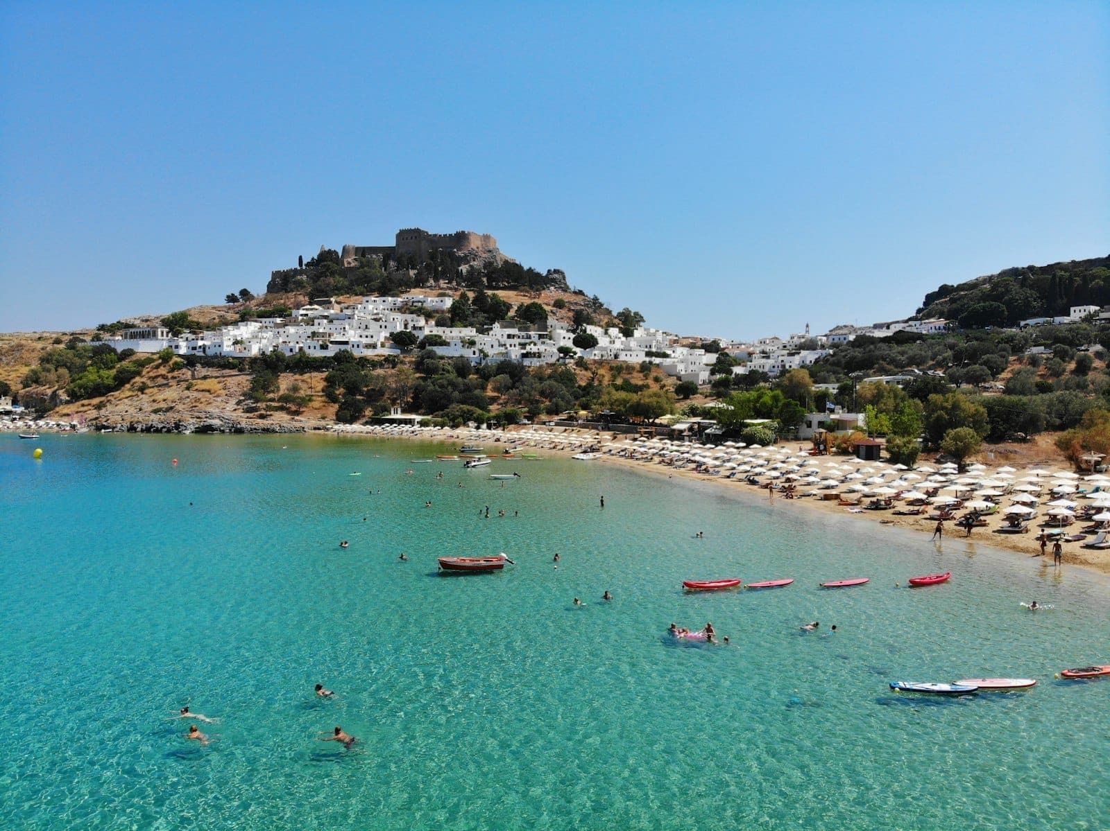 Lindos Beach (Megali Paralia) on Rhodes with crystal-clear turquoise water, sunbathers on sandy shore, and the Acropolis of Lindos above whitewashed houses.