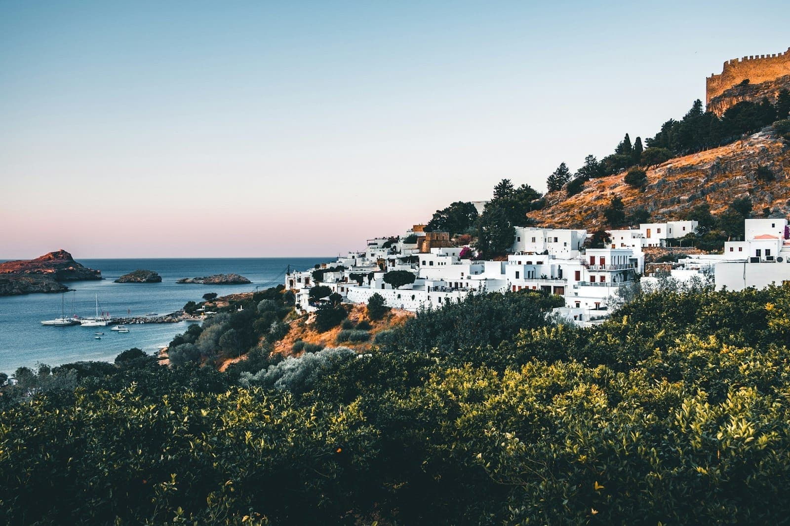 View of Lindos village with whitewashed houses nestled along a hillside, the ancient acropolis above, and the sea in the background at sunset.