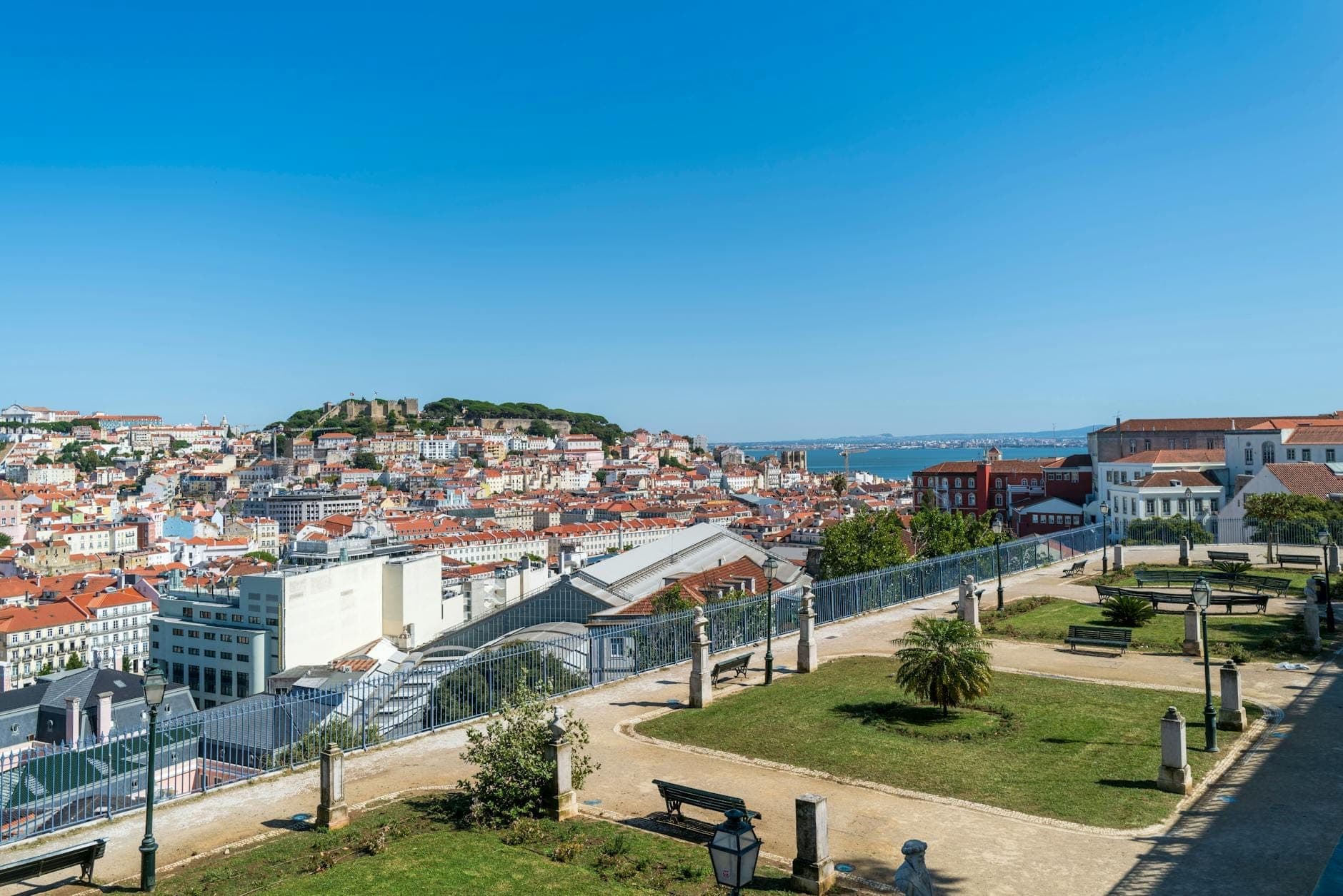Miradouro de terraço aberto com vista para a cidade de Lisboa, com bancos e vistas panorâmicas de edifícios com telhados vermelhos e o rio sob um céu azul limpo.