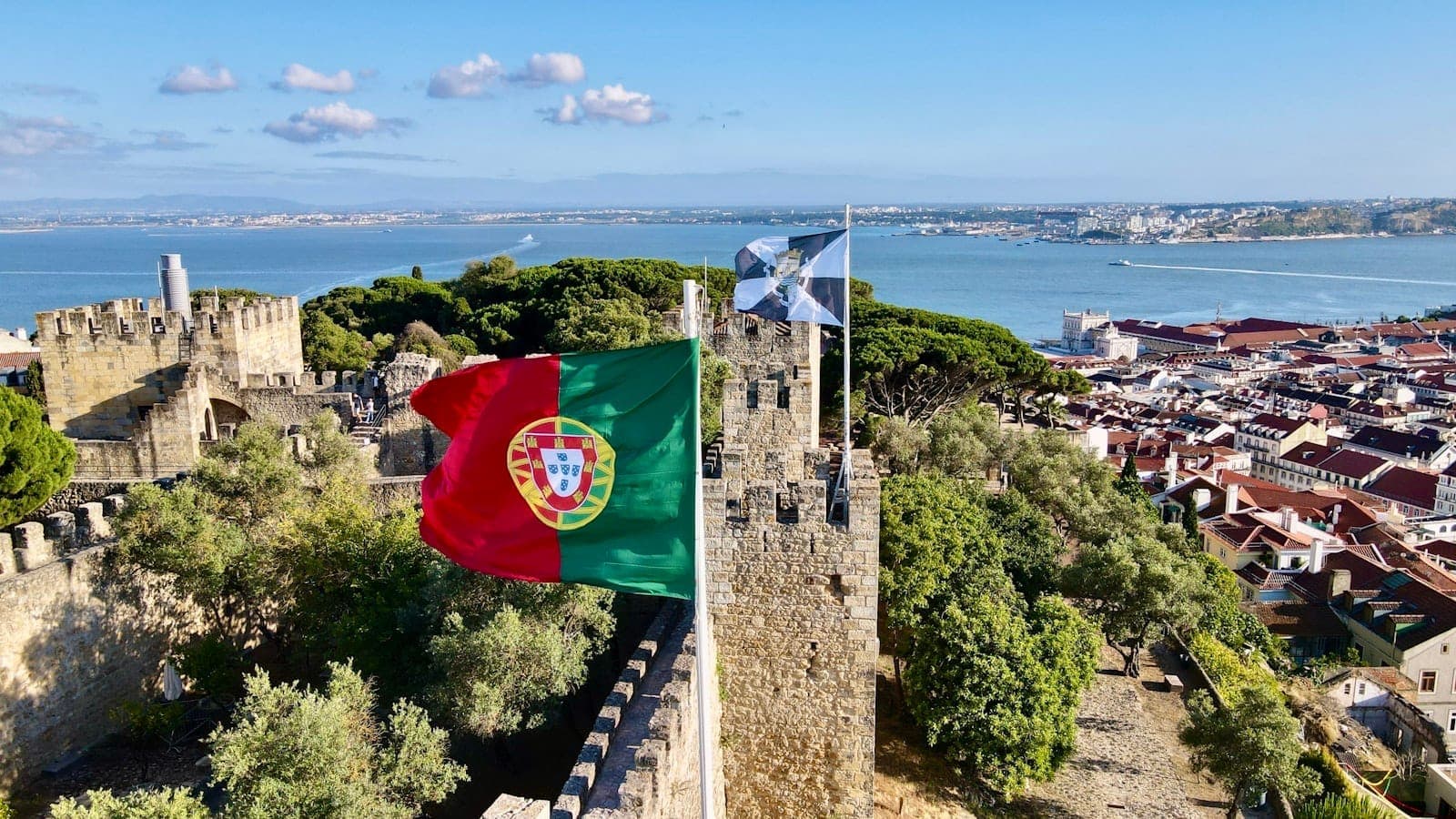Vista das muralhas do Castelo de São Jorge com uma bandeira portuguesa, o rio Tejo e os telhados de Lisboa sob um céu azul.
