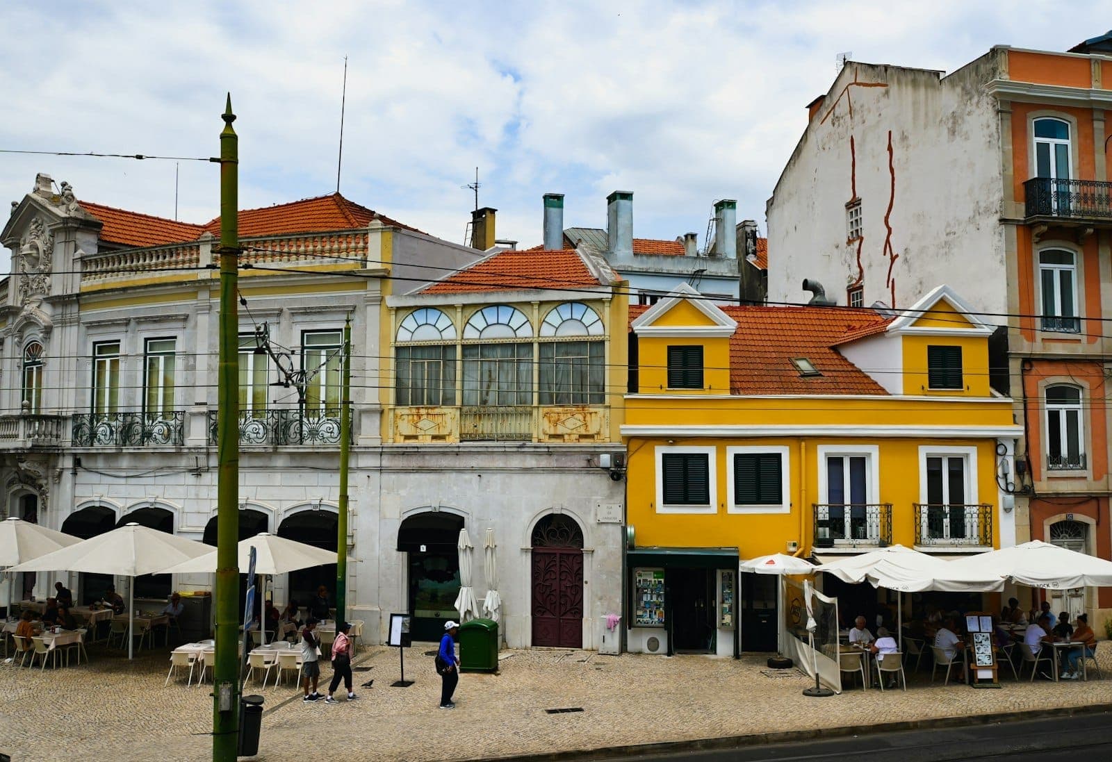 Fileira de edifícios coloridos de Lisboa com vários cafés ao ar livre e pessoas a jantar sob guarda-sóis num dia ensolarado.