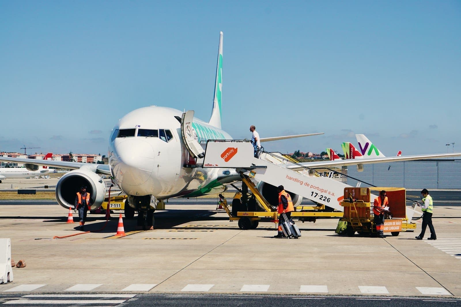 Vista rente ao solo de um avião comercial no Aeroporto de Lisboa, com passageiros embarcando pela escada e funcionários trabalhando na pista.