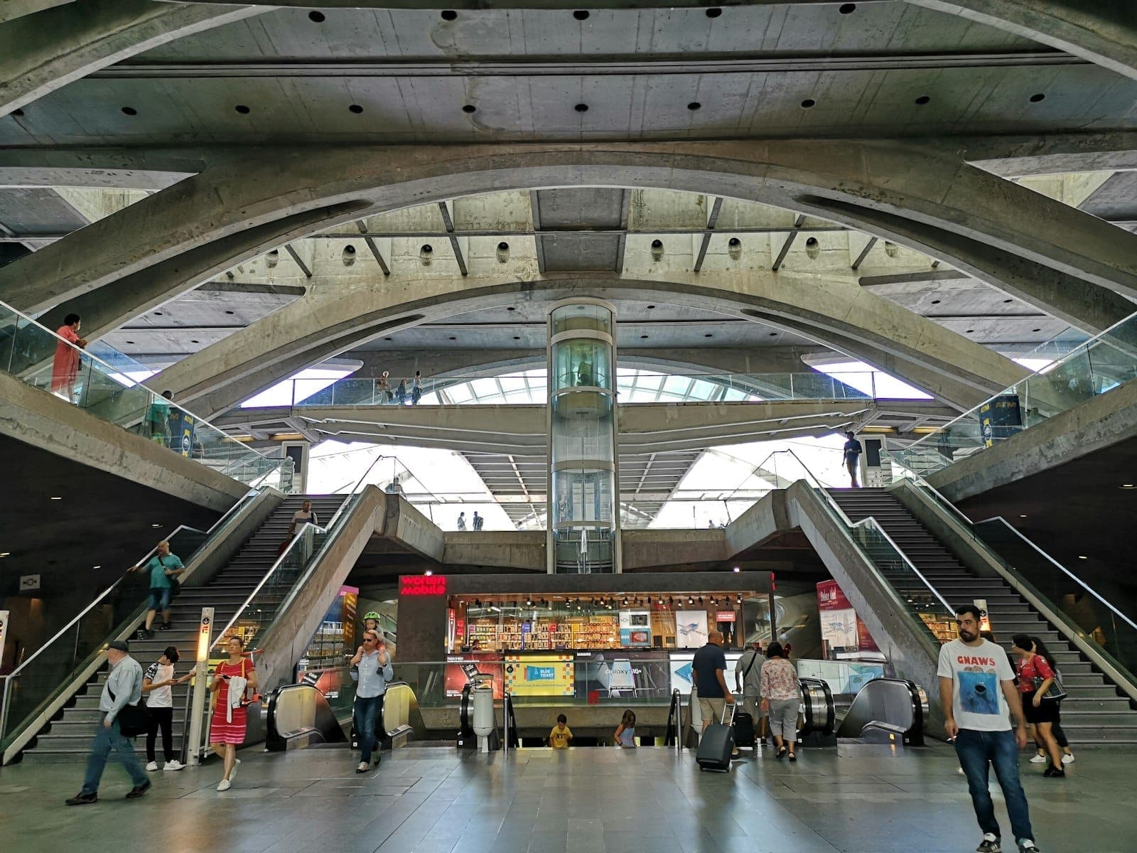 Vista interior do Aeroporto de Lisboa com passageiros, escadas rolantes e arquitetura moderna em betão, mostrando as instalações do aeroporto e a atividade na área de chegadas.