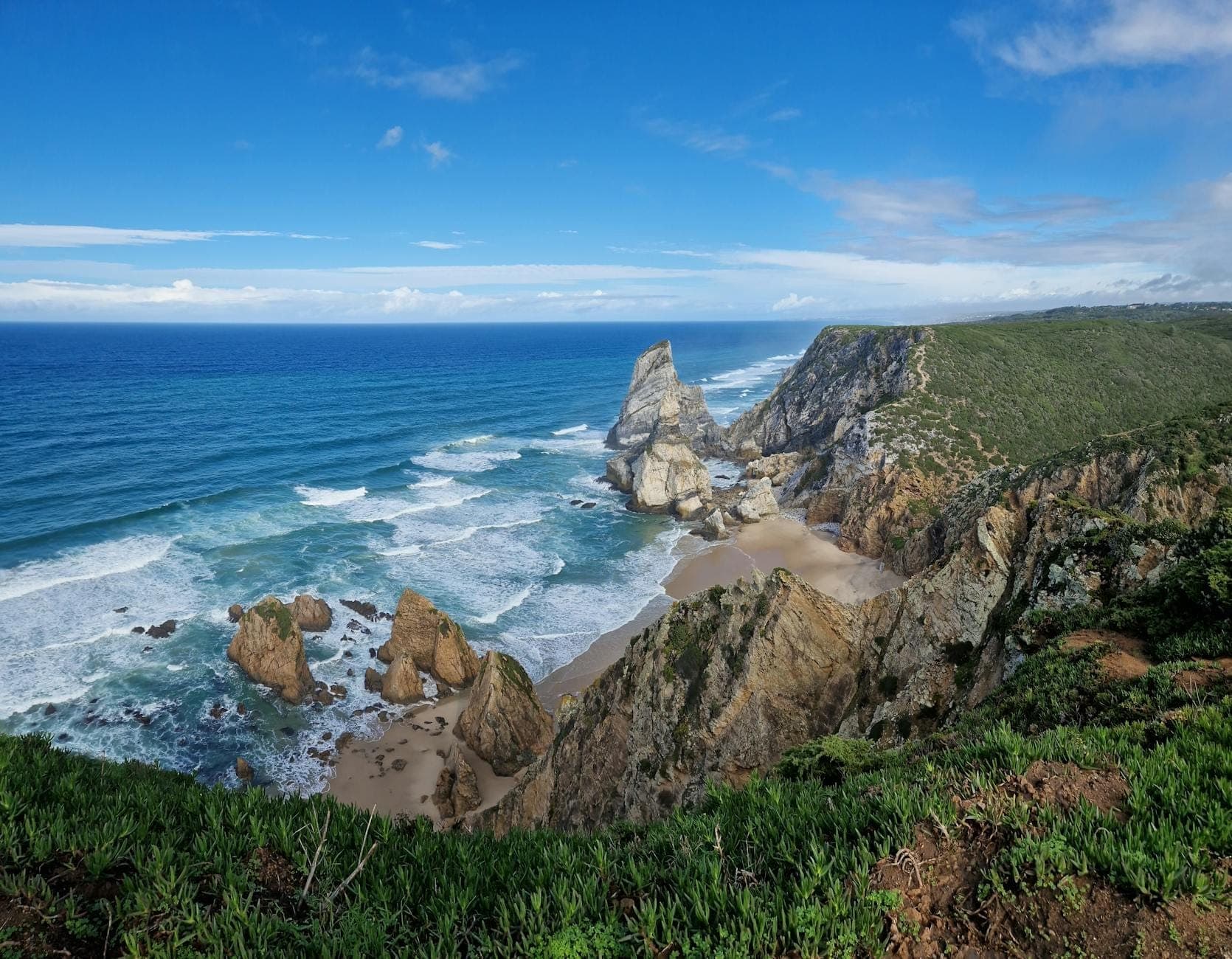 Falésias dramáticas e praia atlântica selvagem com formações rochosas e ondas a oeste de Cascais sob um céu azul.