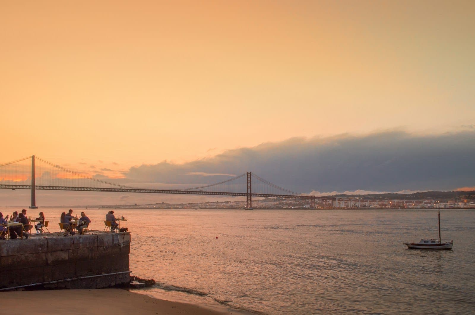Pessoas sentadas numa esplanada à beira-rio ao pôr do sol com a Ponte 25 de Abril e um veleiro ao fundo em Lisboa.