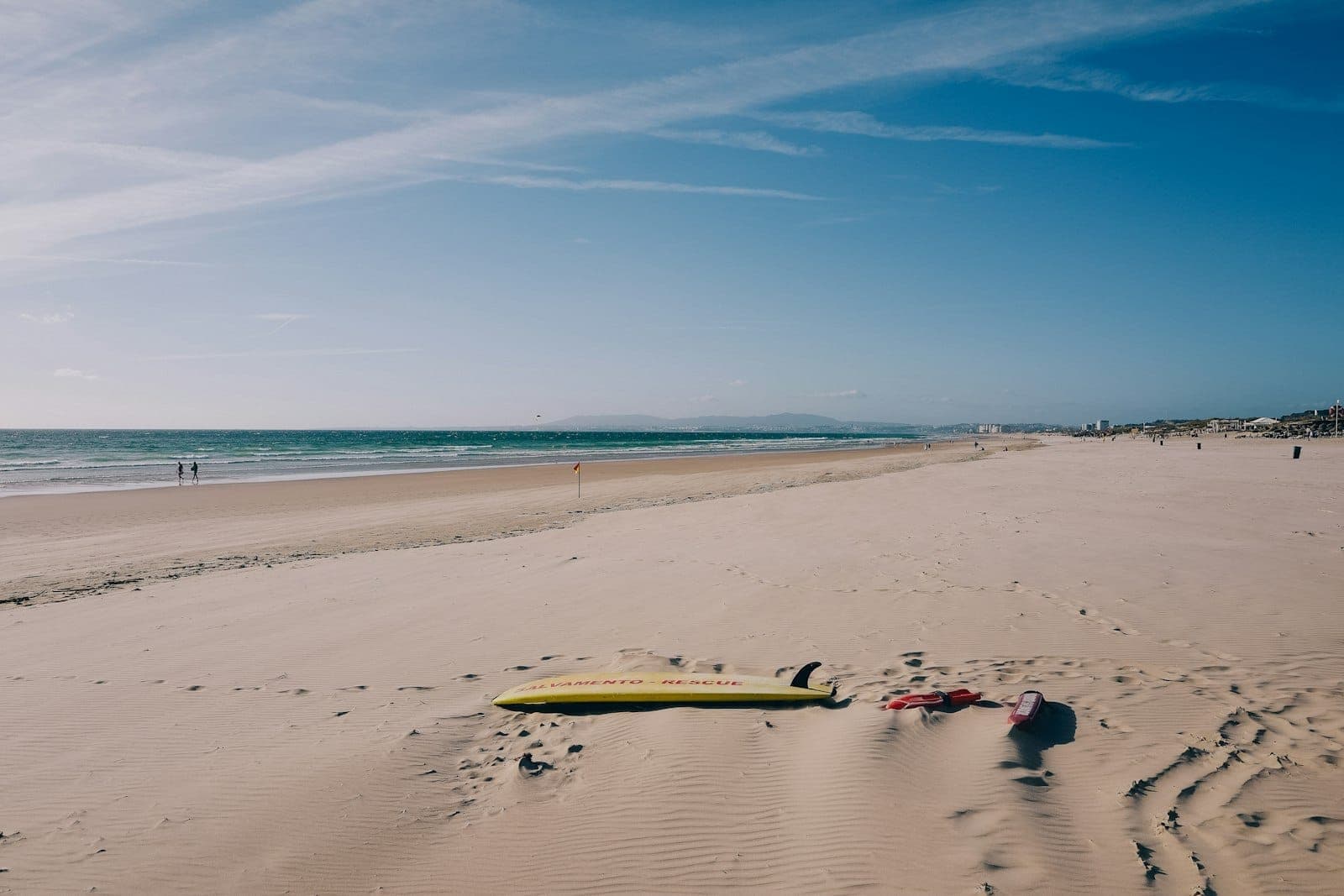 Ampla praia de areia perto de Lisboa com uma prancha de surfe na areia e céu azul, transmitindo relaxamento e aventura na costa atlântica.