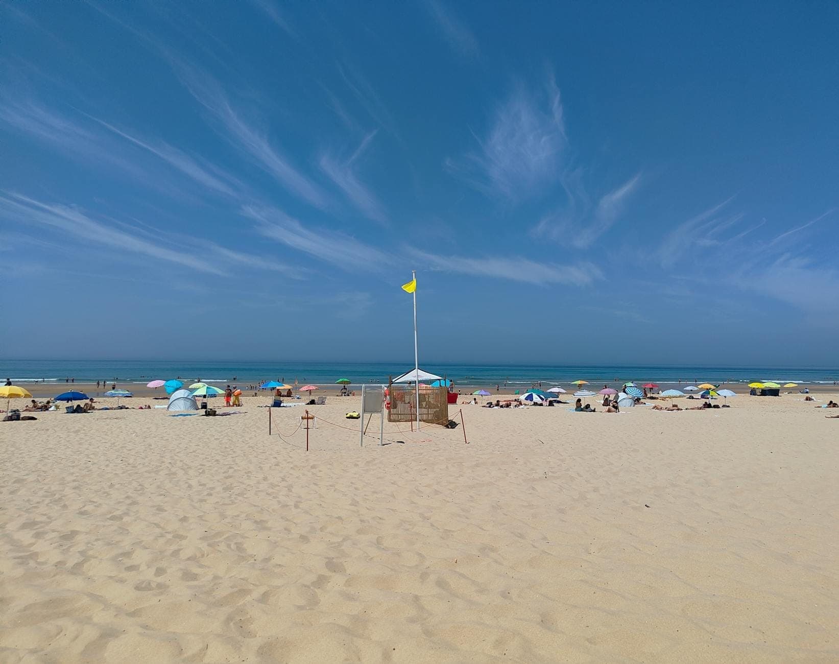 Ampla cena de praia de areia com guarda-sóis coloridos e banhistas sob um céu azul limpo, de frente para o Atlântico perto de Lisboa.