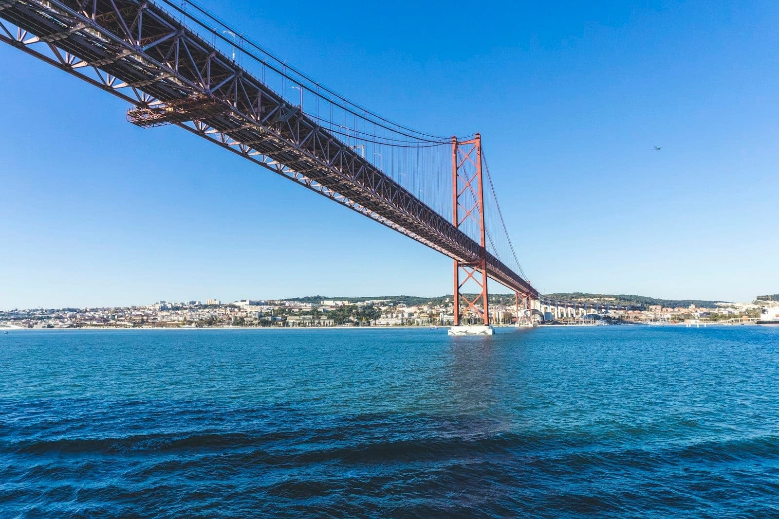 Vista dramática em ângulo baixo da Ponte 25 de Abril em Lisboa mostrando o pilone sul e a água abaixo em um dia claro.