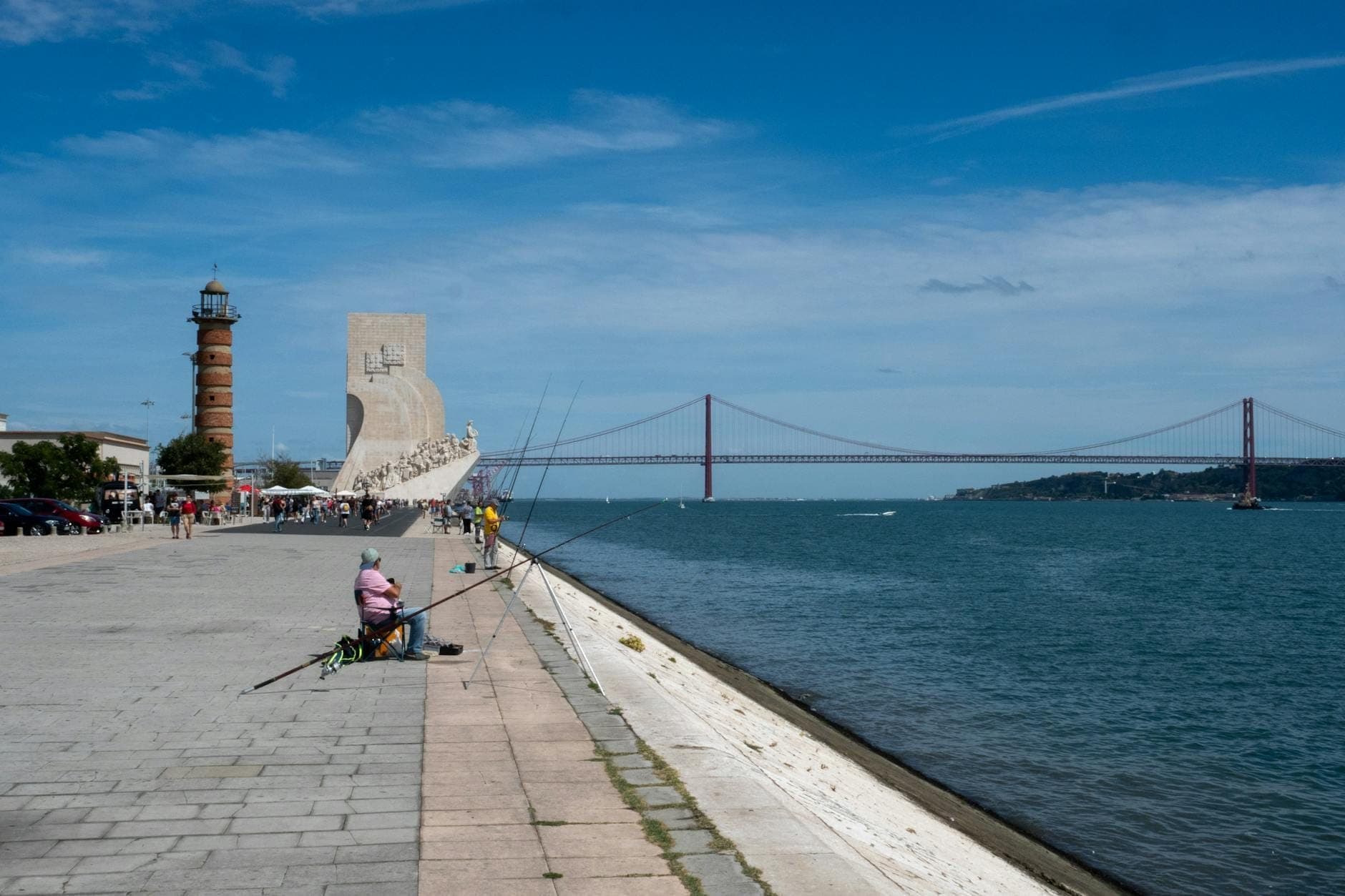 Orla marítima de Lisboa em Belém com a Ponte 25 de Abril, um farol e o Padrão dos Descobrimentos visíveis sob um céu azul.
