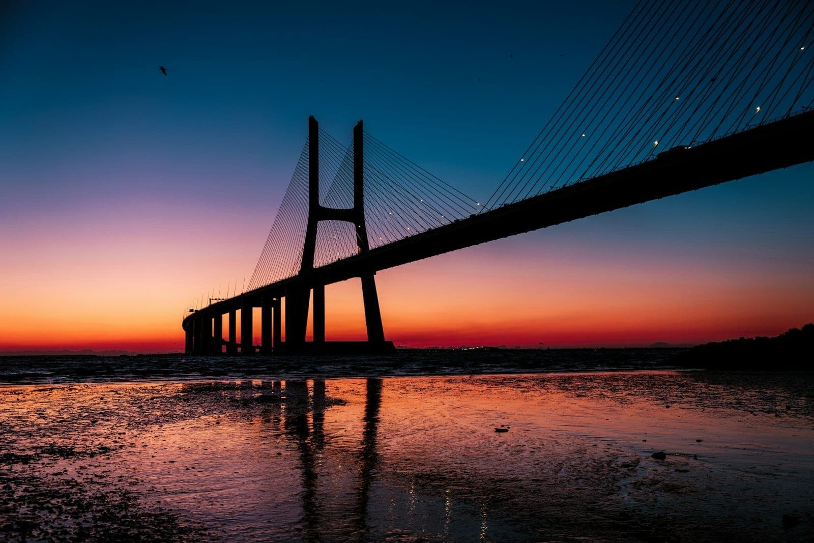 Ponte Vasco da Gama estendendo-se sobre o rio Tejo ao amanhecer, com silhuetas dramáticas e reflexos na margem molhada do rio.