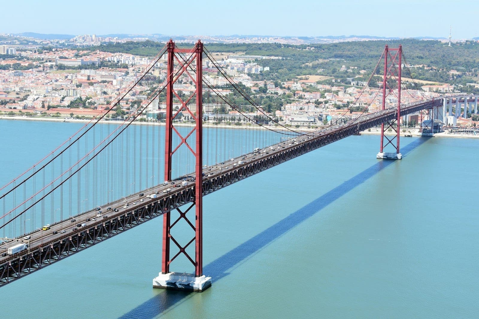 Vista aérea ampla da Ponte 25 de Abril de Lisboa com suas icônicas torres vermelhas cruzando o rio Tejo em frente à paisagem urbana.