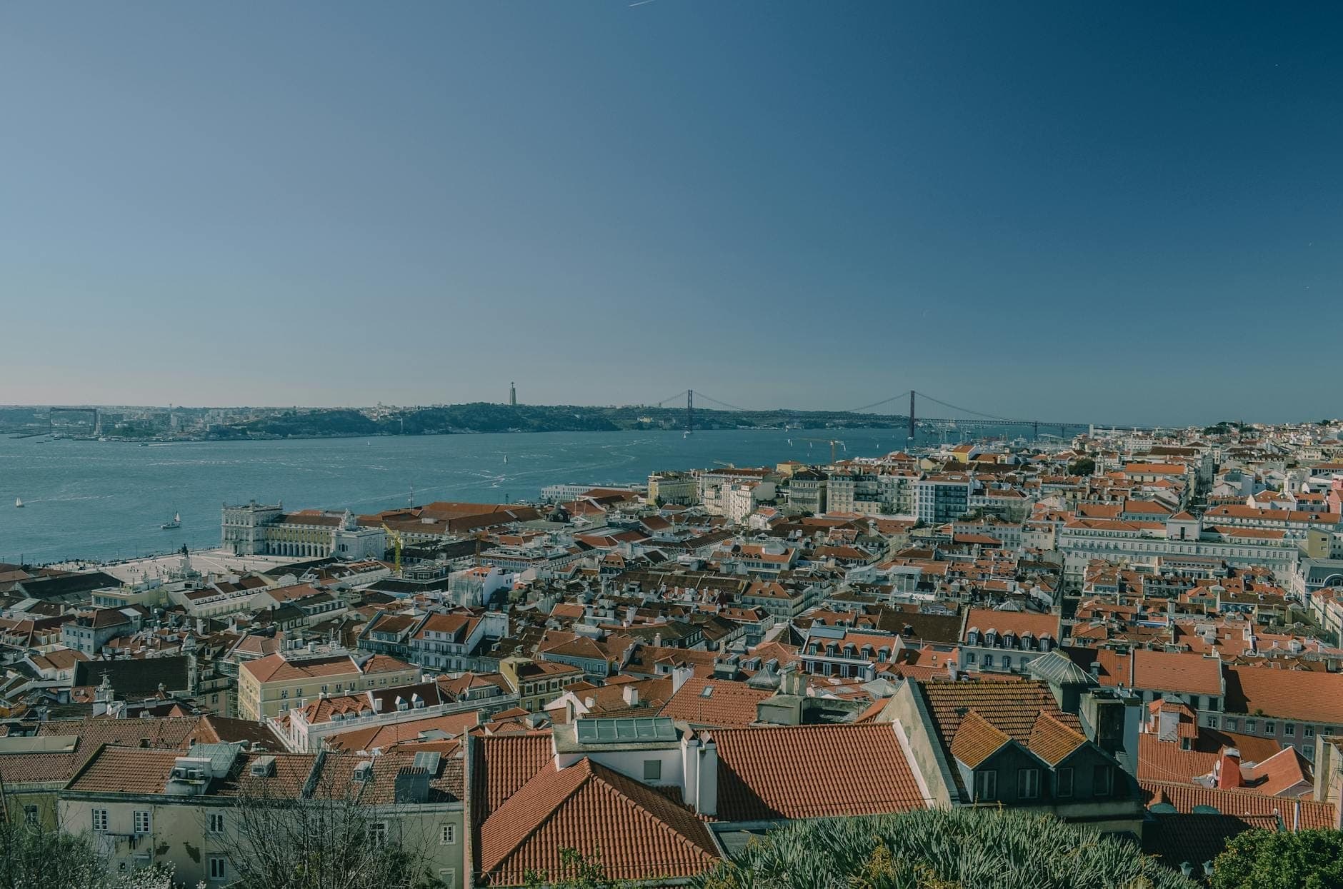 Vista ampla sobre os telhados vermelhos de Lisboa com o rio Tejo e a Ponte 25 de Abril visíveis ao fundo sob um céu azul limpo.