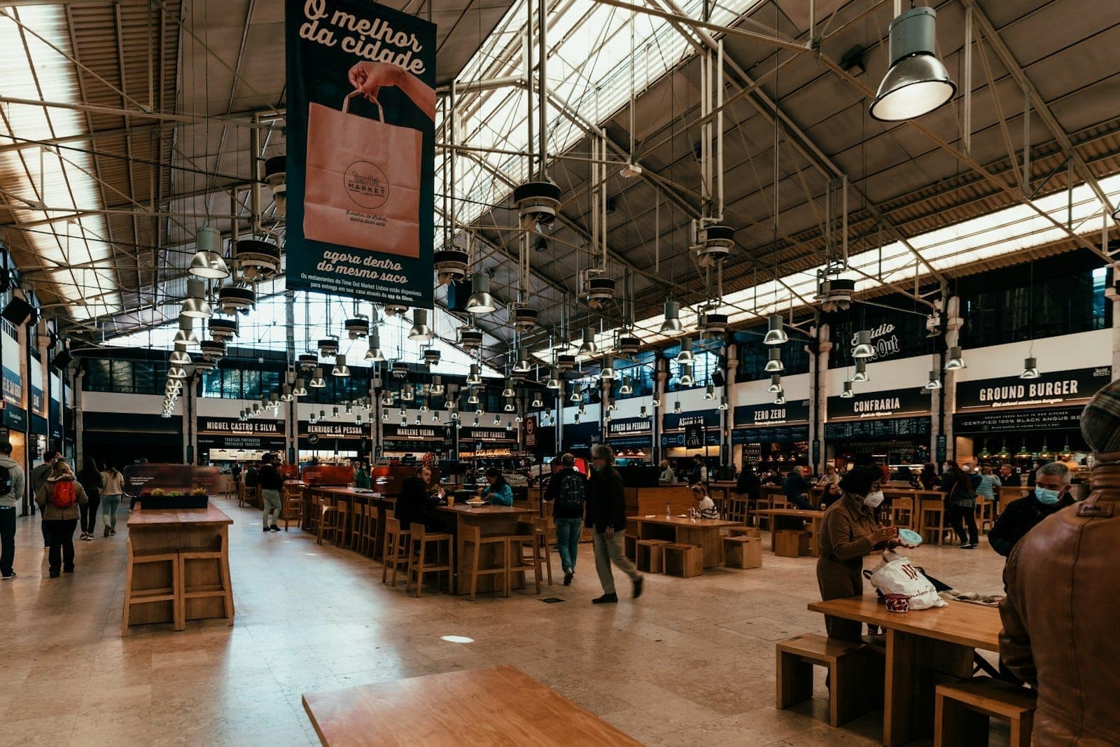 Interior do Mercado da Ribeira em Lisboa com mesas comunitárias, bancas de comida e pessoas a comer.