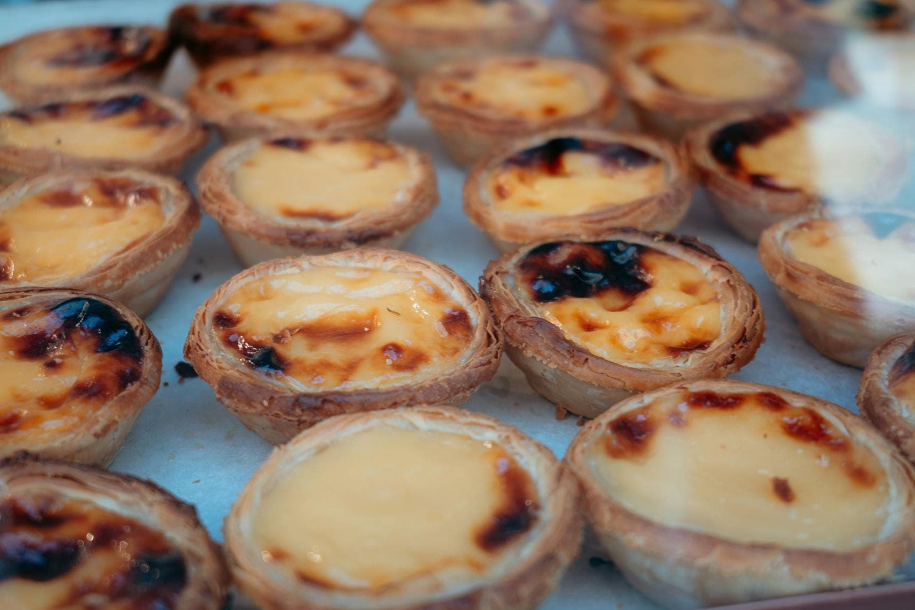 Uma grande bandeja de pastéis de nata frescos, dourados e ligeiramente caramelizados por cima, expostos numa padaria.