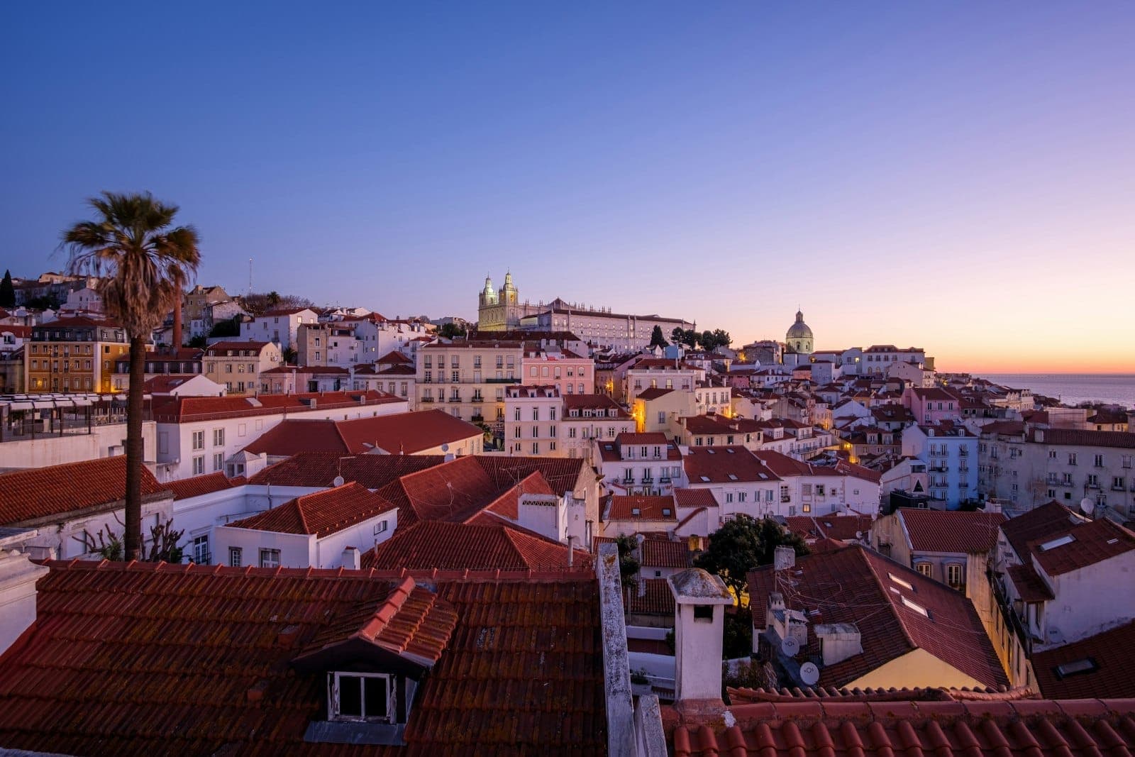Vista noturna dos telhados de Lisboa com um céu azul profundo e luzes quentes, capturando uma atmosfera romântica ao entardecer.
