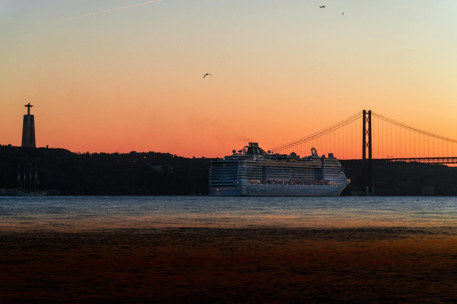 Um grande navio de cruzeiro no rio Tejo ao pôr do sol com a Ponte 25 de Abril e a estátua do Cristo Rei ao fundo.