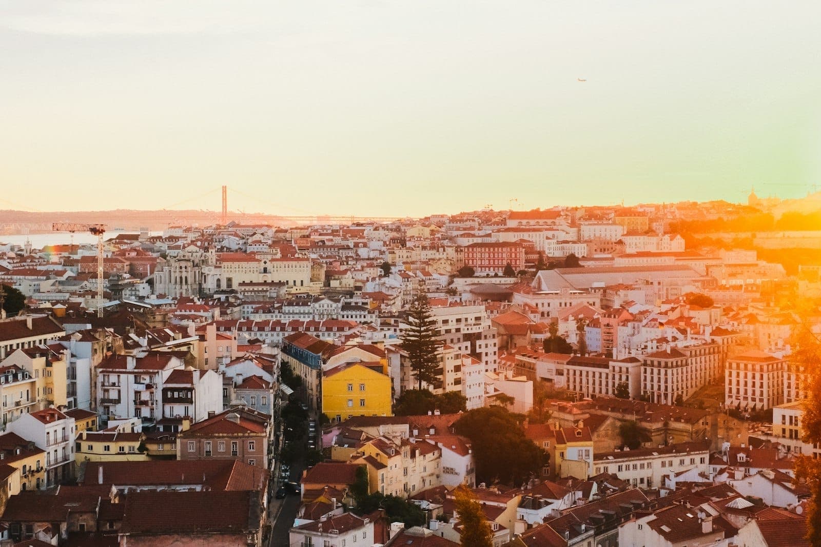 Panorama de Lisboa ao pôr do sol, com telhados de terracota, edifícios coloridos e uma suave luz dourada iluminando o horizonte e os marcos distantes.