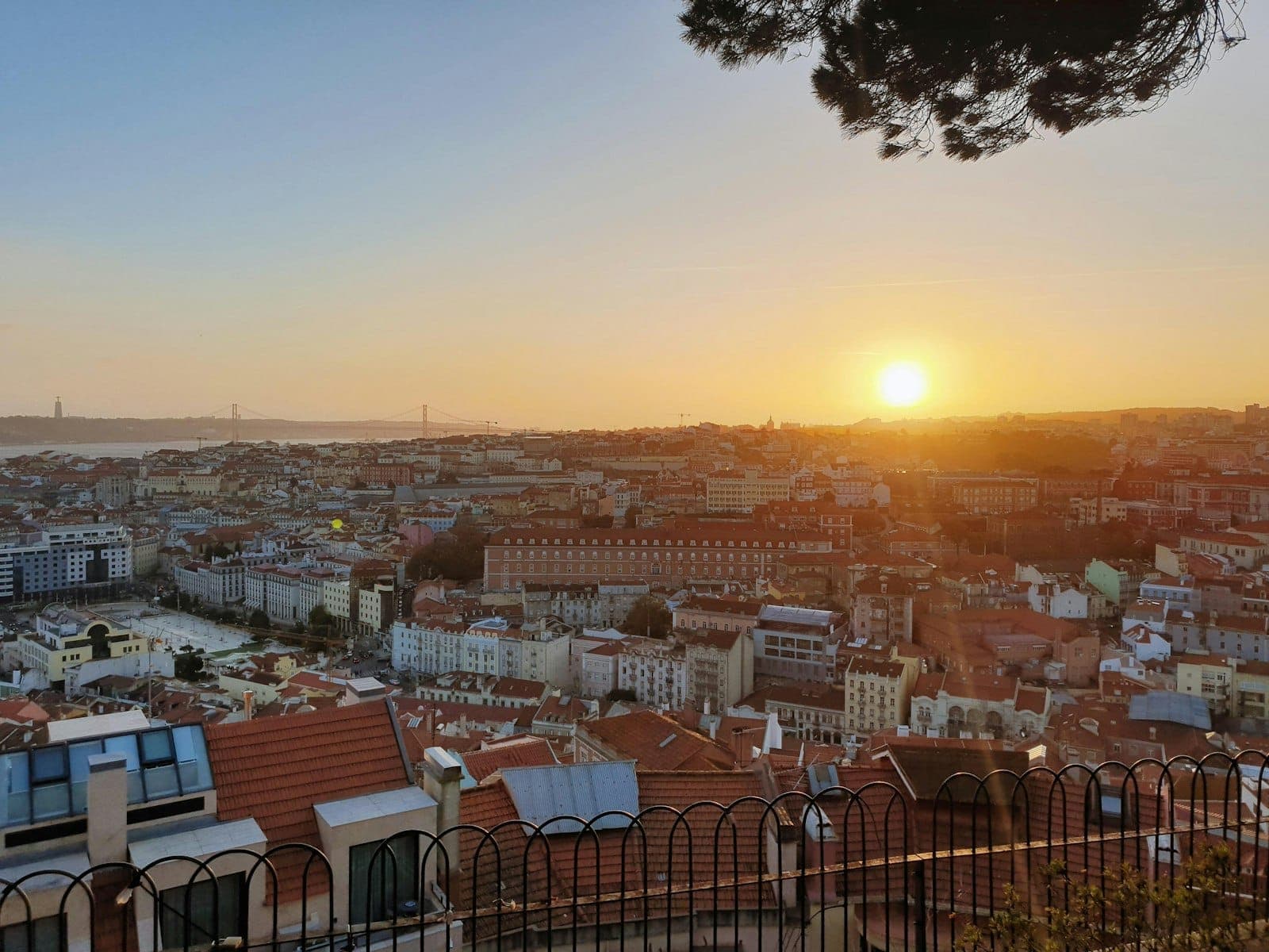 Vista do pôr do sol sobre os telhados de Lisboa e o rio Tejo, vista de um miradouro elevado com uma grade de ferro e galhos de árvores acima.