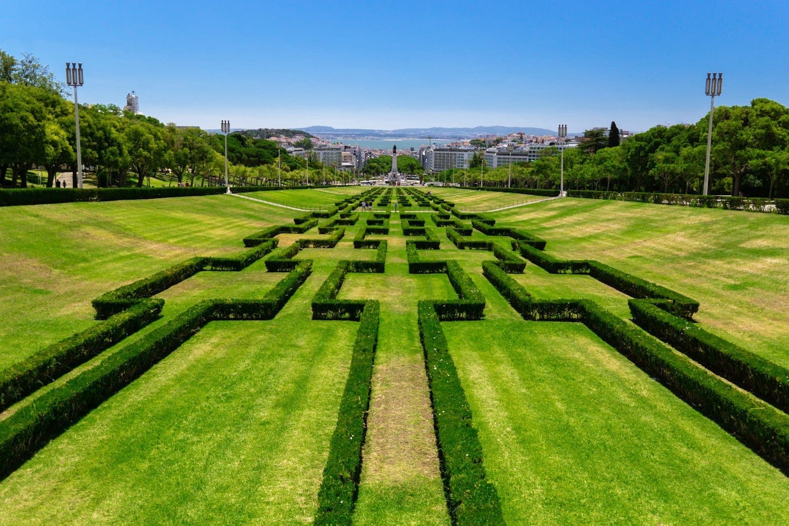 Jardins simétricos e exuberantes com sebes aparadas no Parque Eduardo VII de Lisboa num dia ensolarado, com vistas da cidade ao fundo.